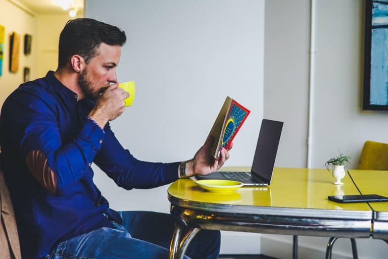 Man drinking coffee reading a book by his laptop